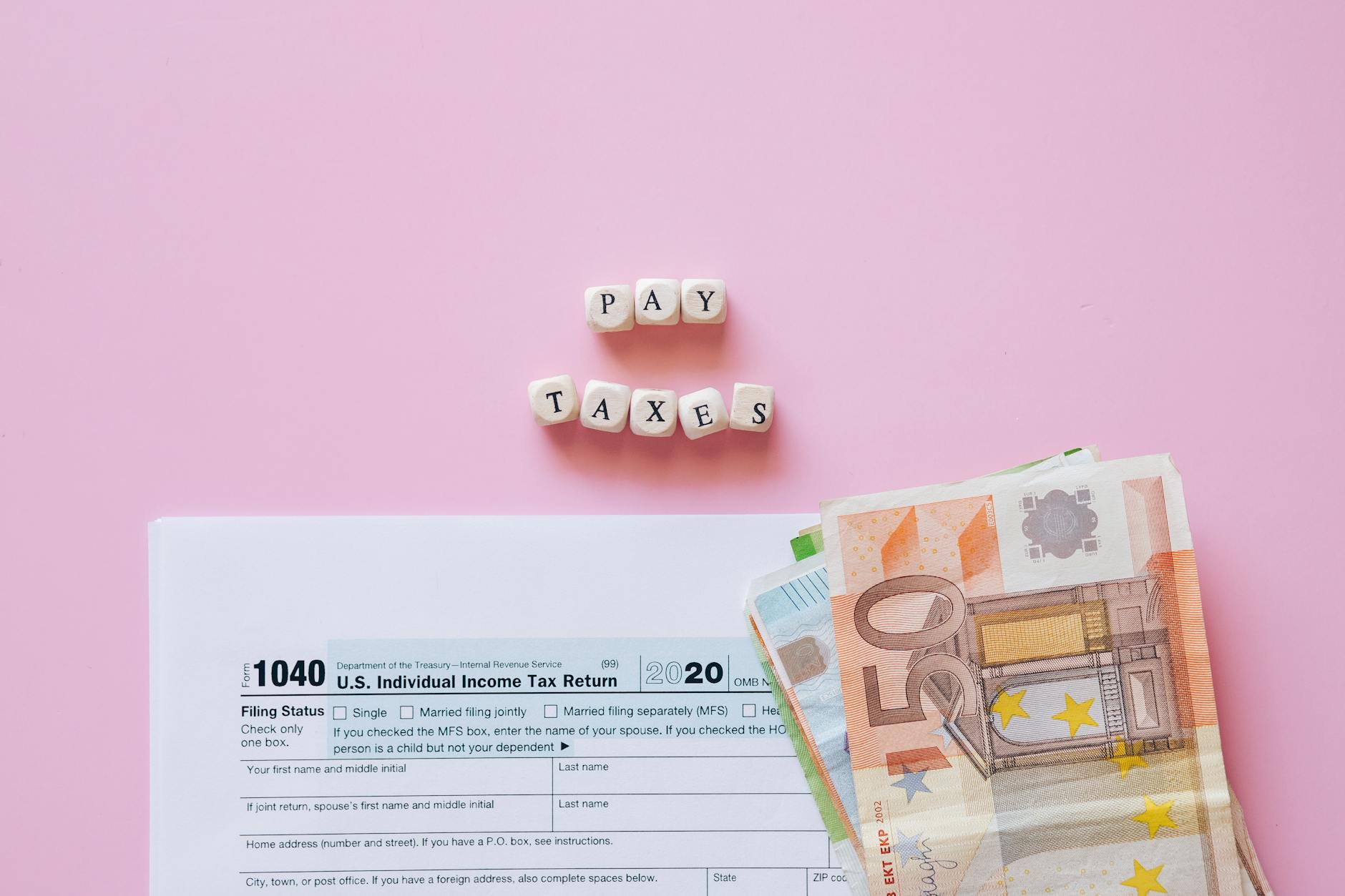 Top view of tax form, euro banknotes, and 'Pay Taxes' letter blocks on pink background.